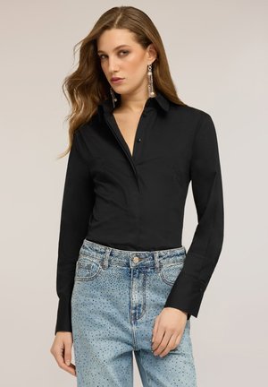 Frau mit langen, welligen Haaren, die ein langärmliges, schwarzes Hemd mit Knöpfen und hellblaue Jeans mit kleinen dunklen Punkten trägt, blickt leicht zur Seite.