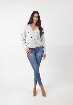Grijze gebreide cardigan met zwarte hartpatronen, v-hals en drie knopen, gecombineerd met blauwe skinny jeans en doorzichtige hakken.