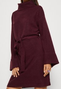 Burgundy stickad klänning med hög polokrage, långa ärmar, bälte i midjan och ribbad textur, med breda manschetter och en avslappnad passform.