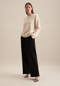 Beige trui met geribde manchetten, gecombineerd met hoge zwarte wijde broek. Model staat in zwarte hoge hakken tegen een neutrale achtergrond.