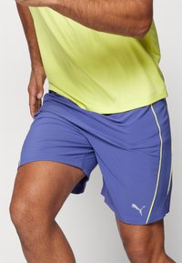Lilla spordipüksid, millel on kerge kangas, kollased küljeaktsendid ja Puma logo alumises servas. Sobib sporditegevuseks.