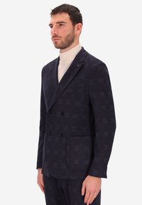 Blazer doppiopetto in tartan navy realizzato in tessuto testurizzato, con bottoni neri e tasche a toppa, indossato sopra un leggero dolcevita.