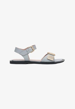 Sandale en cuir gris avec sangle de cheville réglable, semelle plate noire et détail doré à l'avant. Texture lisse et design minimaliste.