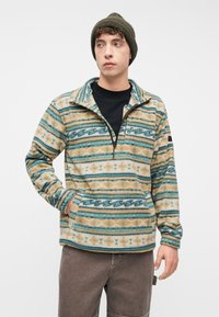 Νέος άνδρας με φλις πουλόβερ με σχέδιο και φερμουάρ στο ένα τέταρτο, μαύρο πουκάμισο, ελια πράσινο σκουφάκι και καφέ παντελόνι, στέκεται με το ένα χέρι στην τσέπη.