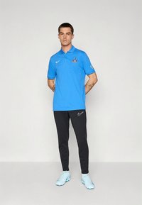 Blaues Nike-Poloshirt mit Logo, schwarze Nike-Hose und hellblaue Fußballschuhe. Das Modell steht aufrecht mit locker an den Seiten hängenden Armen. Einfaches, modernes Design.