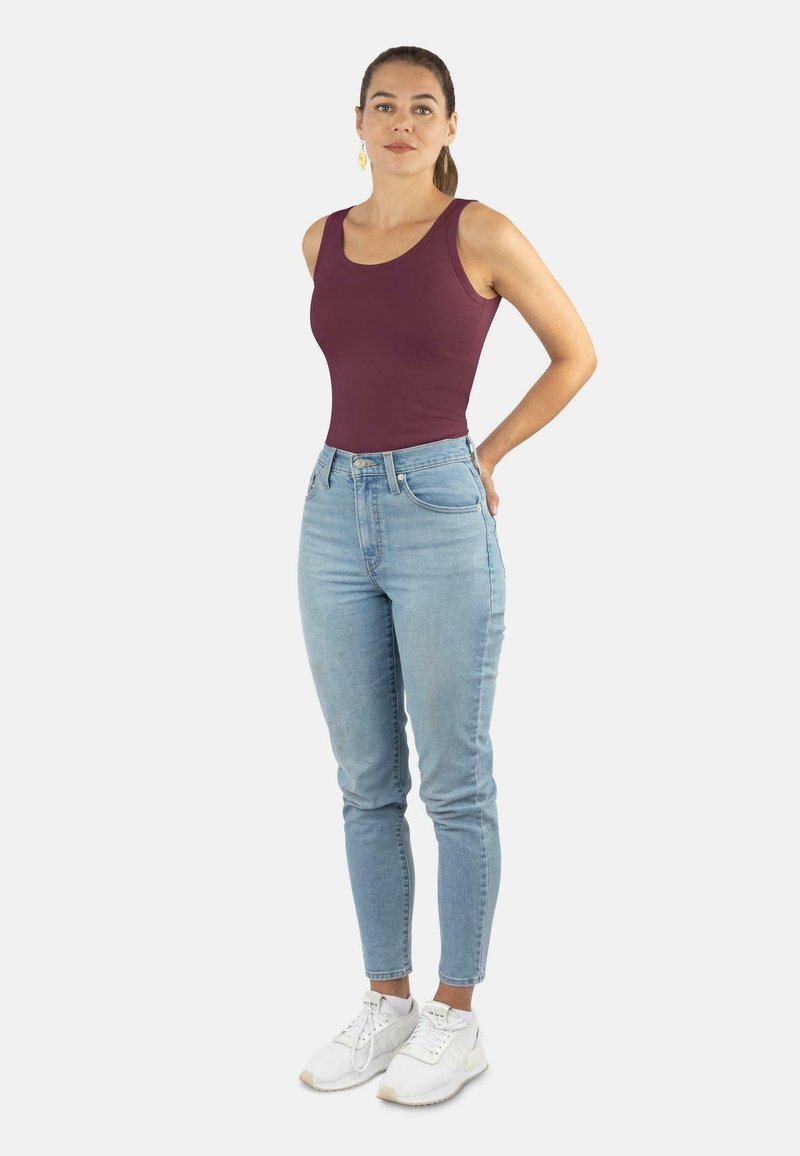 Bordeauxfarbener Tank-Top, hellblaue High-Waist-Jeans, weiße Sneaker. Model steht mit auf Hüften gestützten Händen. Glattes Material, figurbetontes Design, lässiger Stil.