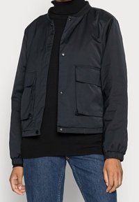 Czarna, lekka kurtka bomber z elastycznymi mankietami, zatrzaskami i dwoma dużymi przednimi kieszeniami, zestawiona z golfem i dżinsami.