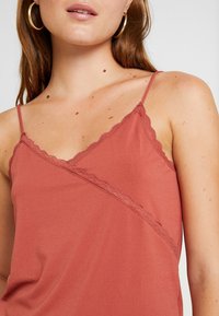 Camisola color ruggine con spalline sottili, caratterizzata da un tessuto testurizzato e bordi in pizzo ondulato lungo il colletto e l'orlo.