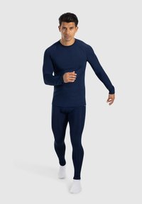 Maglietta sportiva a maniche lunghe blu navy e leggings, con una texture liscia. Il modello è in movimento, indossando calze bianche.