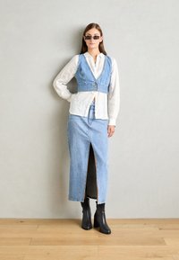 Denim vest with a deep V-neck, button closure, paired with a long denim skirt featuring a front slit. Black ankle boots and sunglasses included.