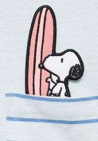 Světle modré tričko z látky má přišité vyobrazení Snoopyho, který drží růžovou surfovací desku, s pruhem detailu kapsy níže.