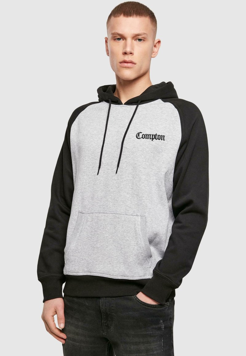 Junger Mann trägt einen grauen und schwarzen Kapuzenpullover mit dem Schriftzug "Compton" auf der Brust und steht mit einer Hand in der Tasche vor schlichtem Hintergrund.