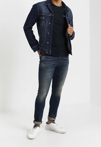 Chaqueta de denim en azul oscuro, con dos bolsillos en el pecho y botones plateados. Lleva puesta una camiseta negra, combinada con jeans de un azul claro y zapatillas blancas.