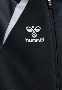 Giacca sportiva nera con zip, caratterizzata da dettagli grigi e un logo "hummel" bianco. Il tessuto ha una finitura strutturata.