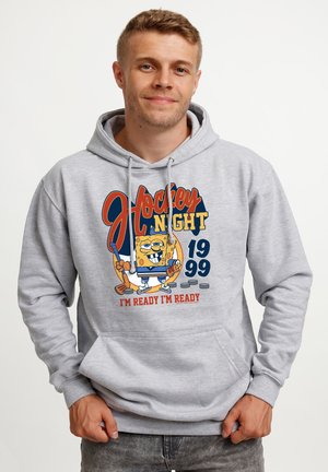 Mężczyzna w szarej bluzie z kapturem z postacią z kreskówki SpongeBob trzymającą kij do hokeja z napisem „Hockey Night 1999 Jestem gotowy Jestem gotowy.”
