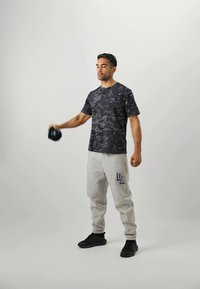 Mænds sort camo kortærmet skjorte i kombination med lysegrå sweatpants med et UA-logo; model holder en håndvægt og har sorte sneakers på.
