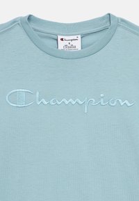 Ανοιχτό μπλε μπλουζάκι Champion με κεντημένο το όνομα της μάρκας στο στήθος και ετικέτα μεγέθους που δείχνει ηλικία 7/8 και μικρό μέγεθος.