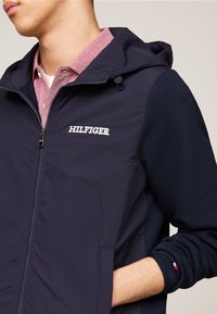 Giacca con zip di colore navy con cappuccio, dotata di maniche navy a contrasto, logo "HILFIGER" e colletto a scacchi rossi sotto.