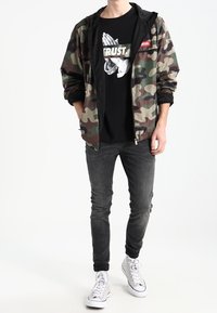 Kamuflaggejakke med mesh-fore, sort grafik t-shirt med teksten "TRUST", slim-fit sorte jeans og hvide sneakers.