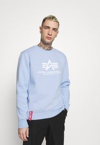 Joven con cabello corto rubio y tatuajes en el cuello, vistiendo un sudadera de Alpha Industries de color azul claro y pantalones negros contra un fondo sencillo.