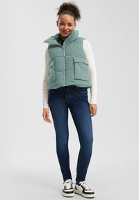 Žena v svetlozelenom prešívanom vestu na bielom tričku s dlhými rukávmi, tmavomodrých úzkych džínsach a čierno-bielych teniskách, stojí a usmieva sa.