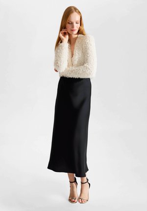 Femme aux longs cheveux roux portant un haut blanc duveteux à manches longues, une jupe midi en satin noire et des talons hauts noirs avec bride à la cheville, debout sur fond blanc.