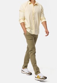 Chemise boutonnée jaune clair, pantalon vert olive et baskets blanches avec des accents gris et jaunes. Coupe ajustée, style décontracté.