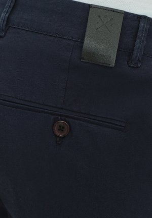 Granatowe spodnie z ciemnobrązowym guzikiem i czarną skórzaną naszywką z geometrycznym wzorem z tyłu. Gładka struktura materiału.