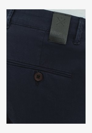 Granatowe spodnie z ciemnobrązowym guzikiem i czarną skórzaną naszywką z geometrycznym wzorem z tyłu. Gładka struktura materiału.