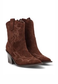 Botines de ante marrón con puntera puntiaguda, costuras de estilo occidental y tacón apilado. Cuentan con lengüetas laterales para facilitar su colocación.