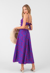 Lila Kleid mit floralem Muster, Spaghetti-Trägern, offenem Rücken und gebundenem Detail. Lässiges Design, fließender Stoff, kombiniert mit einem Strohhut.
