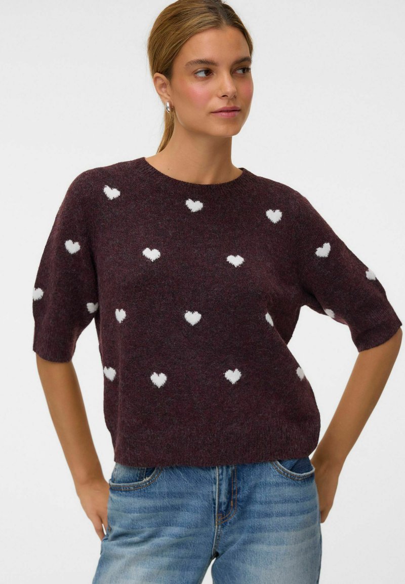 Femme portant un pull court manches violet foncé avec des motifs de cœurs blancs et un jean bleu, debout, mains sur les hanches sur un fond blanc.