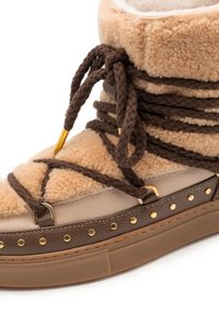 Tan schapenwollen winterlaars met donkerbruine dikke veters eromheen gewikkeld en gouden studs op de leren afwerking.