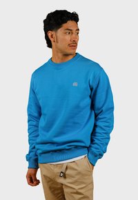 Modrá mikina s ribovaným golierom a man�žetami; obsahuje malé šedé logo slona na hrudi; nosená s béžovými nohavicami.