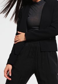 Blazer noir et pantalon plissé noir. Le blazer a une coupe cintrée avec une texture lisse, tandis que le haut est transparent avec un motif subtil.