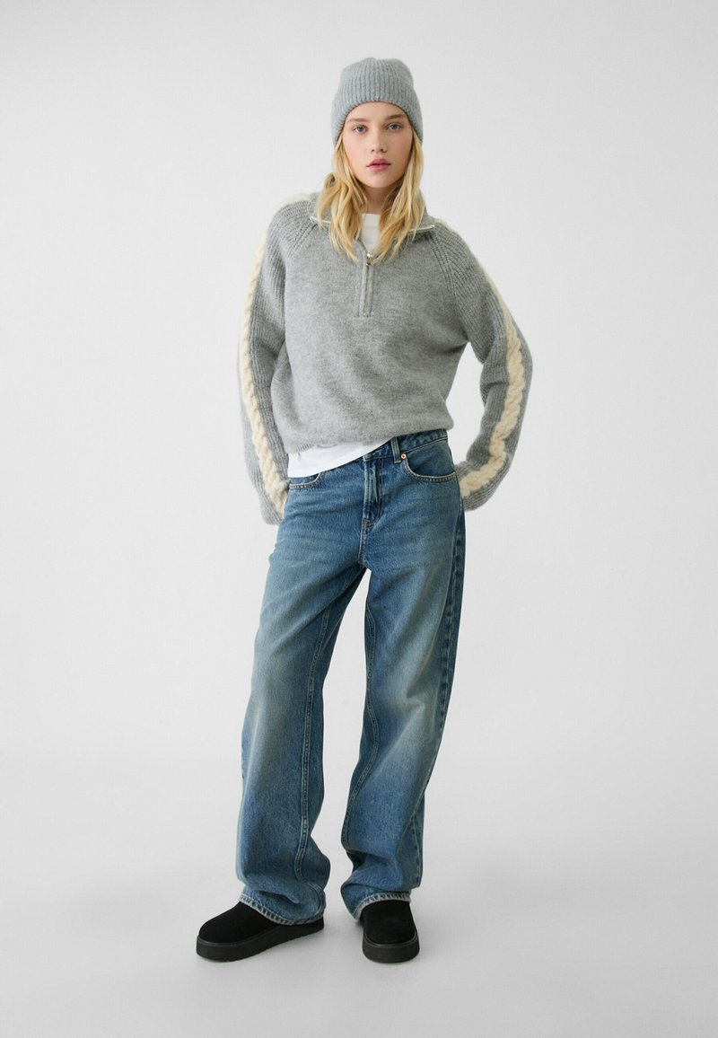 Pullover grigio lavorato a maglia con zip a metà e dettagli intrecciati; abbinato a jeans blu a gamba larga e scarpe nere slip-on. Berretto di maglia grigio in testa.