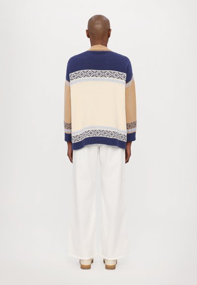 Strickjacke in Marineblau, Creme und Beige mit gemusterten Banddetails. Lässige Passform, gerippte Bündchen und Saum. Getragen über weißen, weiten Hosen.