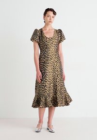 Žena oblečená v predĺžených šatách s leopardím vzorom a nafúknutými rukávmi, so volánovým lemom a striebornými topánkami Mary Jane, stojí pred jednoduchým pozadím.