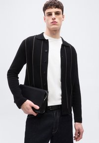 Νεαρός άνδρας που φοράει μαύρη ζακέτα με κουμπιά και λεπτές κάθετες ρίγες, λευκό πουκάμισο, σκούρο τζιν, κρατώντας μια μαύρη τσάντα χειρός με φερμουάρ.