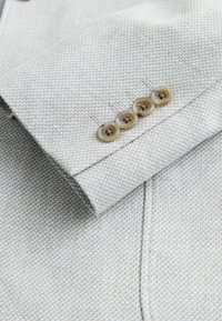 Rukáv svetlosivého textúrovaného obleku so štyrmi svetlohnedými gombíkmi prišitými v rade blízko manžety.