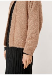 Světle hnědý huňatý cardigan s jemnou texturou a drobným třpytem, otevřený přední design, nošený přes černé plisované šaty s dlouhými rukávy.