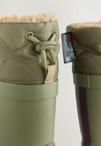 Grüne Gummistiefel mit gesteppter Stoffoberfläche und flauschigem Futter. Enthält einen verstellbaren Riemen und ein Etikett; verfügt über eine glatte und strukturierte Oberfläche.