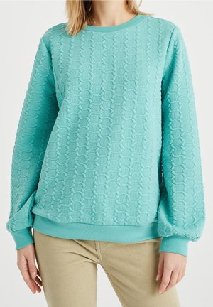 Vrouw met een lange mouw turquoise gestructureerde trui met ballonmouwen en beige broek, staande tegen een effen achtergrond.