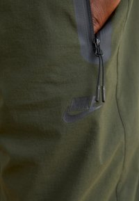 Nike hlače z zadrgo v temno zeleni tkanini z gladko teksturo, ki imajo črn logo in sivo poudarjene detajle ob zadrgi.