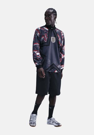 Osoba stojąca w ciemnej, graficznej, długorękawowej koszulce piłkarskiej Anglii, czarnych spodenkach, czarnych skarpetach i srebrnych butach sportowych Nike.