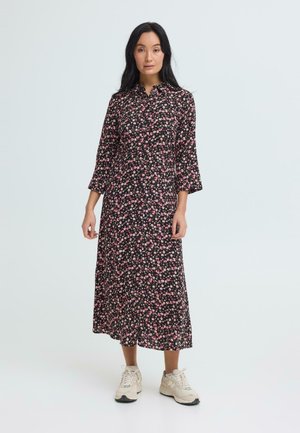 Kobieta stojąca, ubrana w czarną sukienkę midi w różowy kwiatowy wzór oraz białe trampki, na tle jasnego, jednolitego tła.