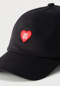 Μαύρο καπέλο μπέιζμπολ με κόκκινο πατς σε σχήμα καρδιάς, κεντημένο με λευκό κείμενο "HUGO" στην μπροστινή όψη.