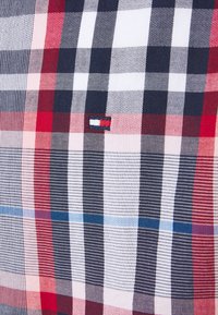 Gros plan sur un tissu à carreaux rouge, blanc, marine et bleu clair avec un petit emblème rectangulaire de drapeau en marine, rouge et blanc cousu au centre.