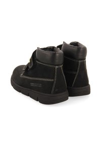 Bottines en suede noir avec un col rembourré, des coutures blanches et une semelle en caoutchouc. Elles comportent une sangle en velcro et un détail de texte en relief.