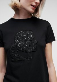 Svart t-shirt med en texturerad svart paljettsapplikation av en profildesign. Korta ärmar, rund hals, slät tyg.